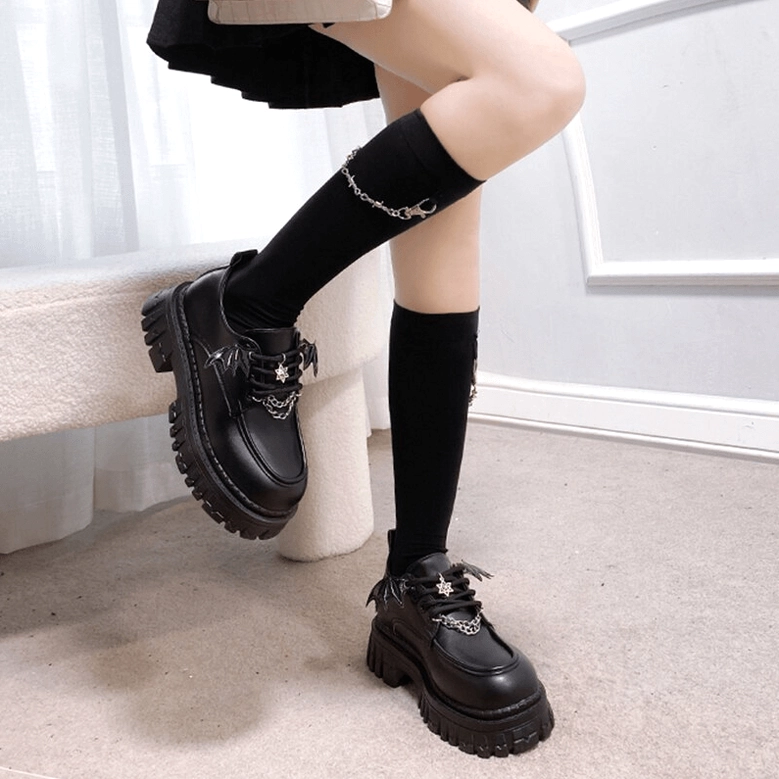 Female model in black platform loafers with bat wing embellishments, wearing black knee-high socks, side angled view on bench