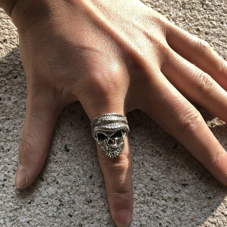 Bearded skull ring worn on male left hand index finger, back top view on textured concrete background showing gothic styling in daily wear