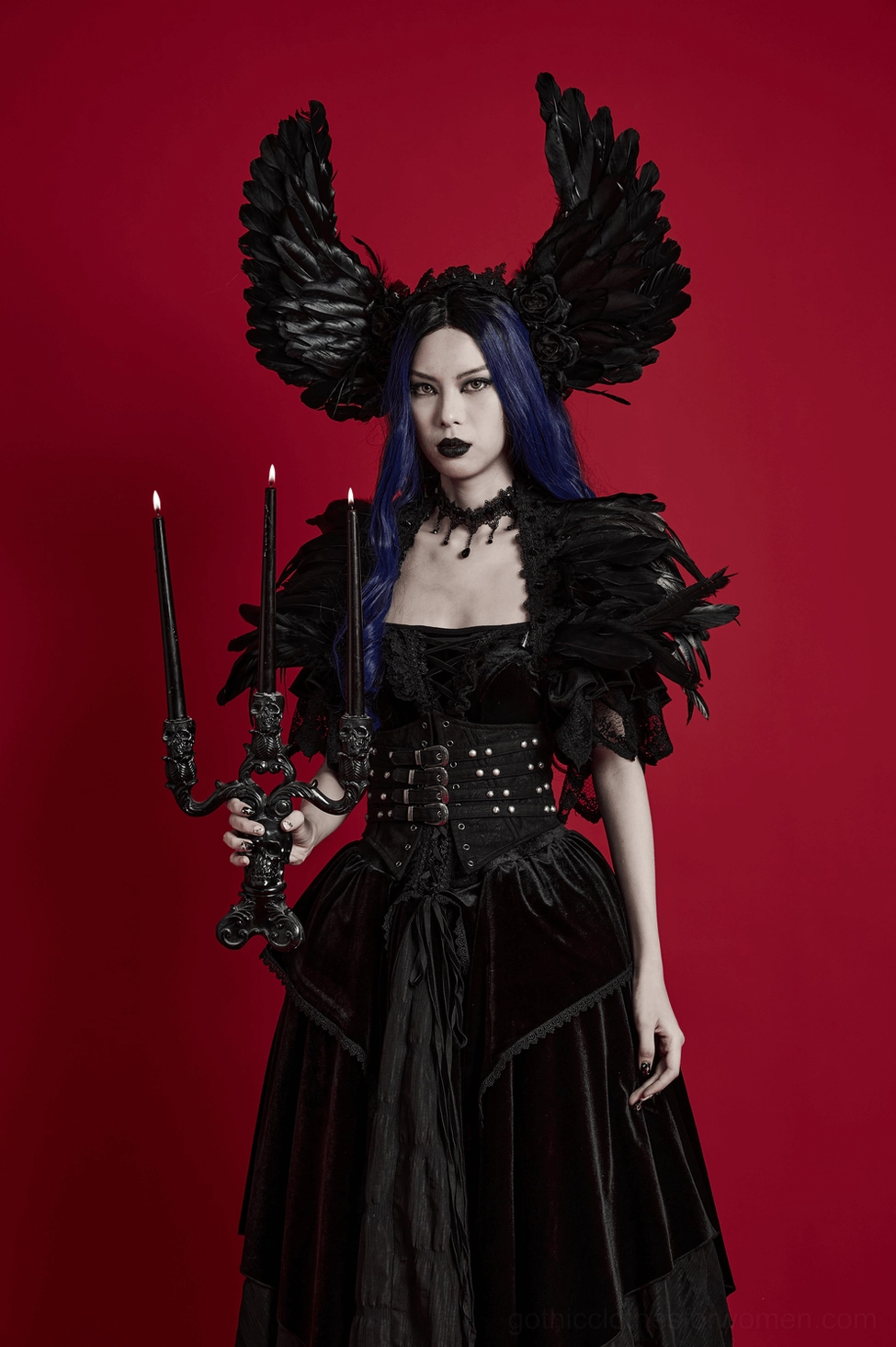Full body styling shot of model wearing black feather devil wings headband with gothic off-shoulder dress and black candlestick prop on red background