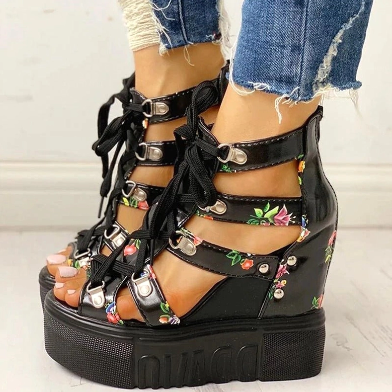 Black floral platform wedge gladiator sandals with multiple buckle straps and lace-up front, front close-up on light wood background