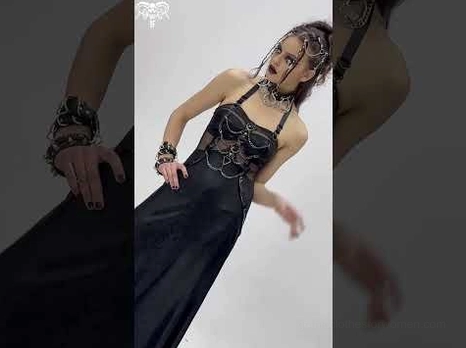 Female goth model in black halter chain maxi dress with skull accessories, side three-quarter pose showing dramatic silhouette