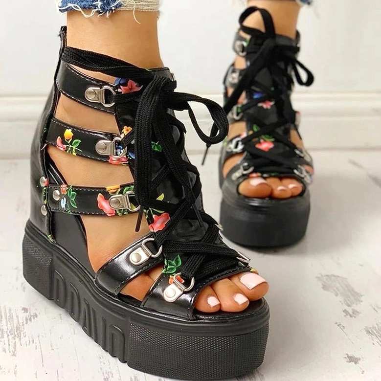 Black floral platform wedge sandals with multiple buckle straps and lace-up details, front side close-up on light wood grain background