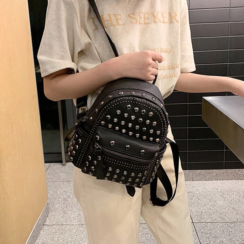 Side front angle of black studded gothic backpack held by shoulder strap showing compact size and pebbled leather texture