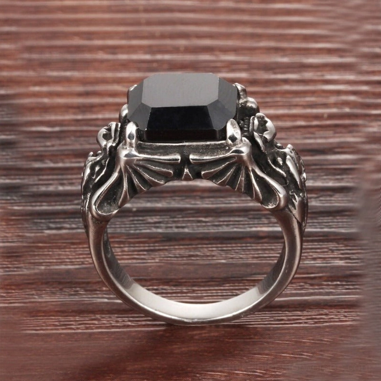 Black zircon gothic statement ring top-down view revealing dark silver tone, winged skull motifs framing square black gem, wood grain surface backdrop