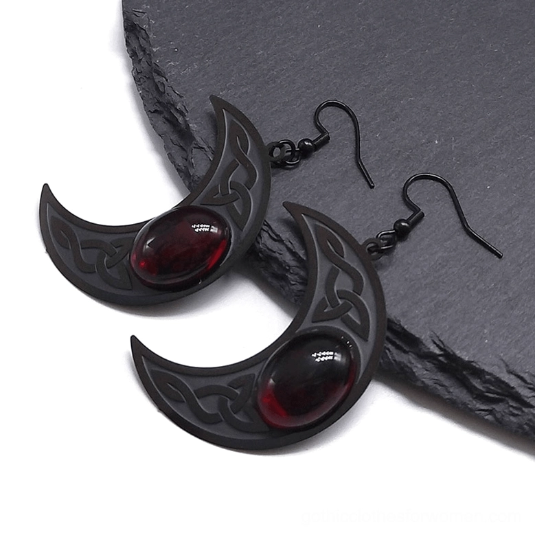 Angled view of Celtic crescent moon earrings with dark red oval gem, matte black metal, engraved details on slate background