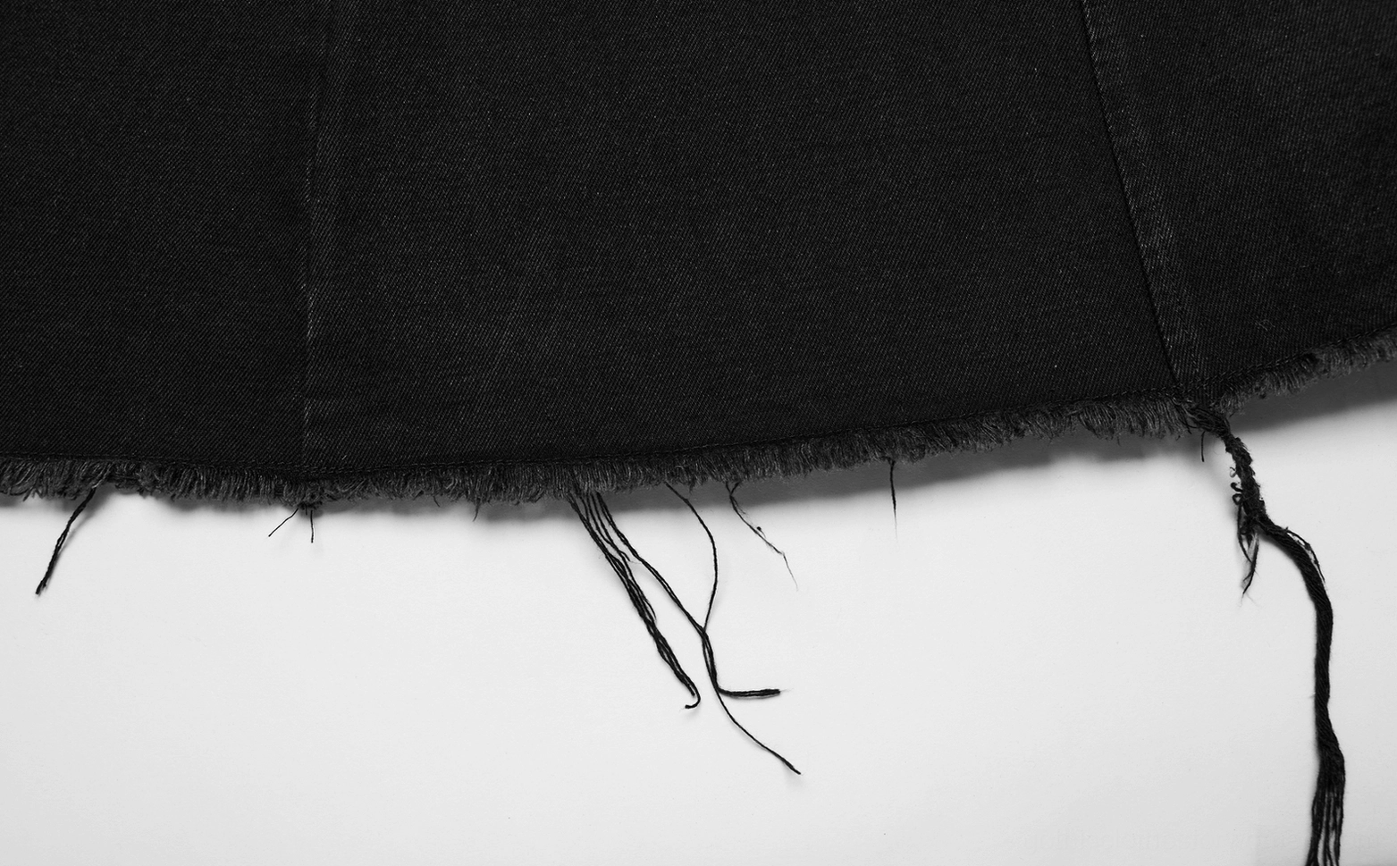 Closeup detail of black denim cape hem, raw frayed edge with loose fabric threads, characteristic deconstructed finish