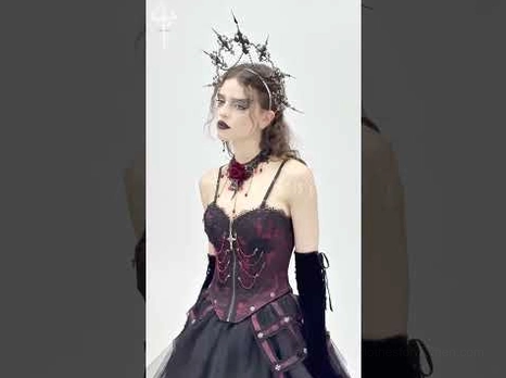 Goth model wearing wine red gothic corset with cross pendant, rose choker, black gloves and crown headpiece, side profile view
