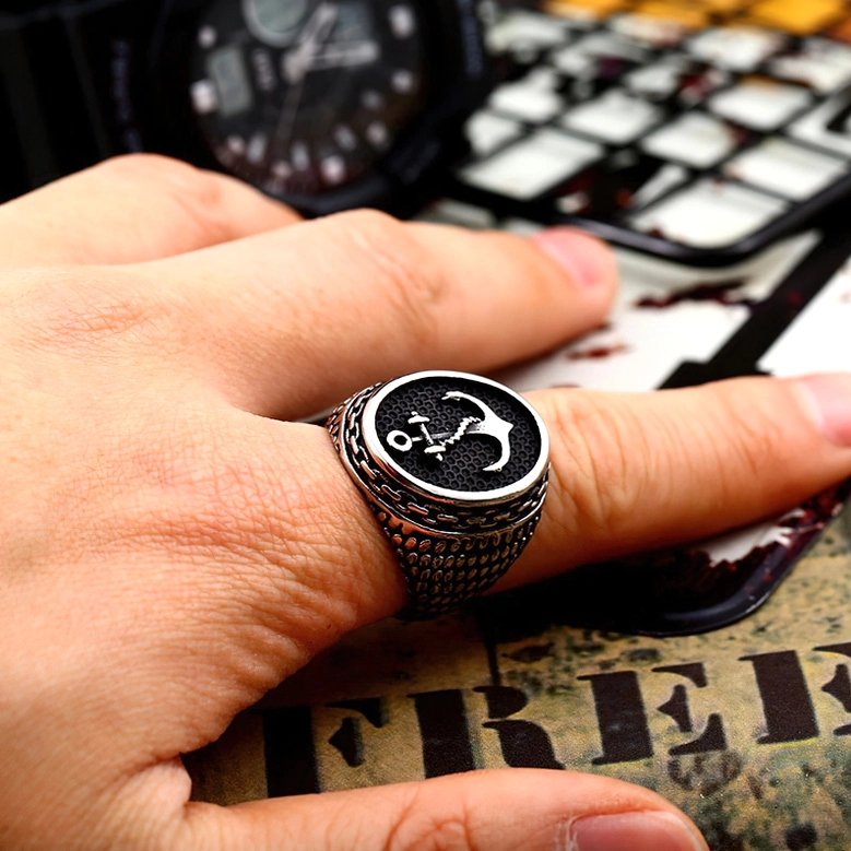 Alternative anchor signet ring side close-up worn on index finger with watch and vintage print backdrop, scale-textured band detail