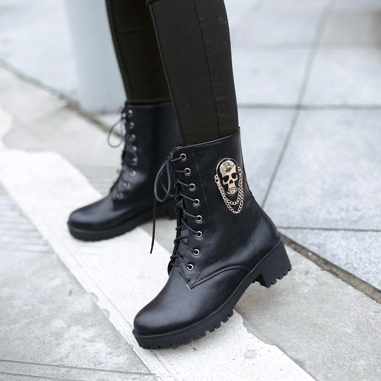 Ground-level side shot of woman wearing black gothic platform ankle boots on tiled street, showing rugged lug sole and skull chain detail