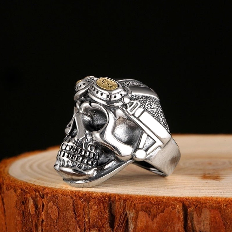 Left three-quarter view of adjustable aviator skull biker ring showing gold-toned goggle lens highlights on wooden display