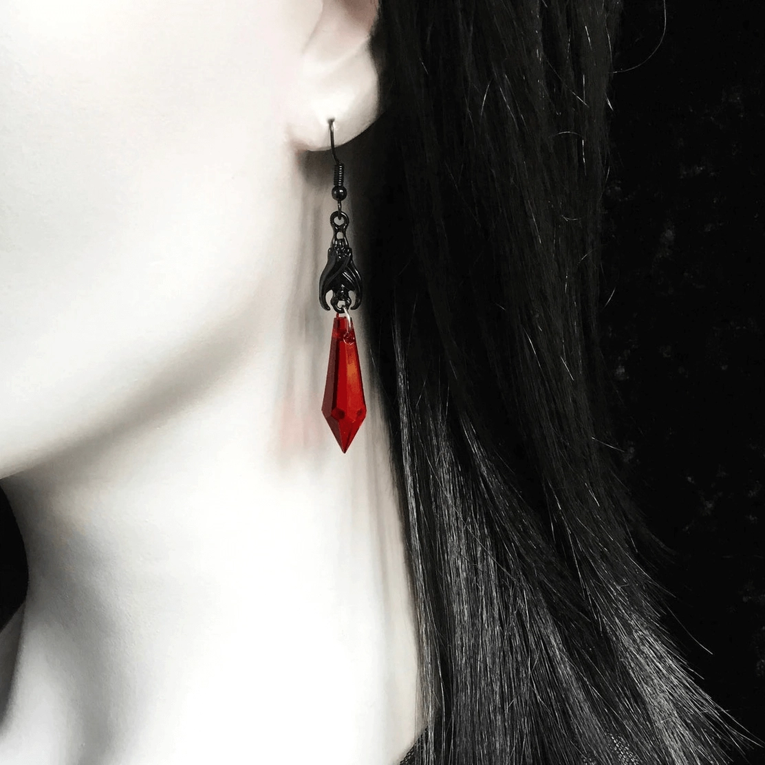 Gothic spike drop earrings worn on female model with long black hair, side close-up view showing red faceted teardrop pendant