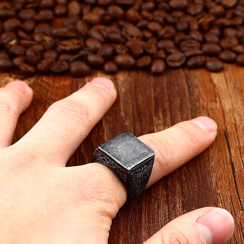 Aged stainless steel gothic square ring worn on index finger, styled on hand against wooden table with coffee beans