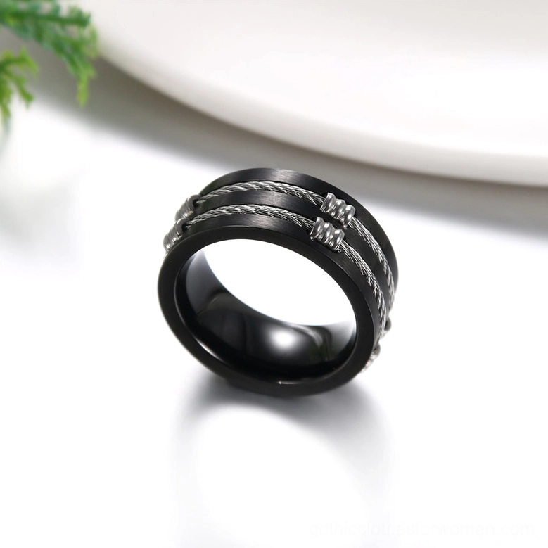 Black stainless steel gothic ring on tabletop with white plate and green plant backdrop, 3/4 oblique view showing smooth glossy finish and silver wire details