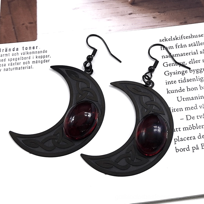 Vintage style flat lay of black crescent moon earrings with red gemstone, Celtic knot engraving on printed paper
