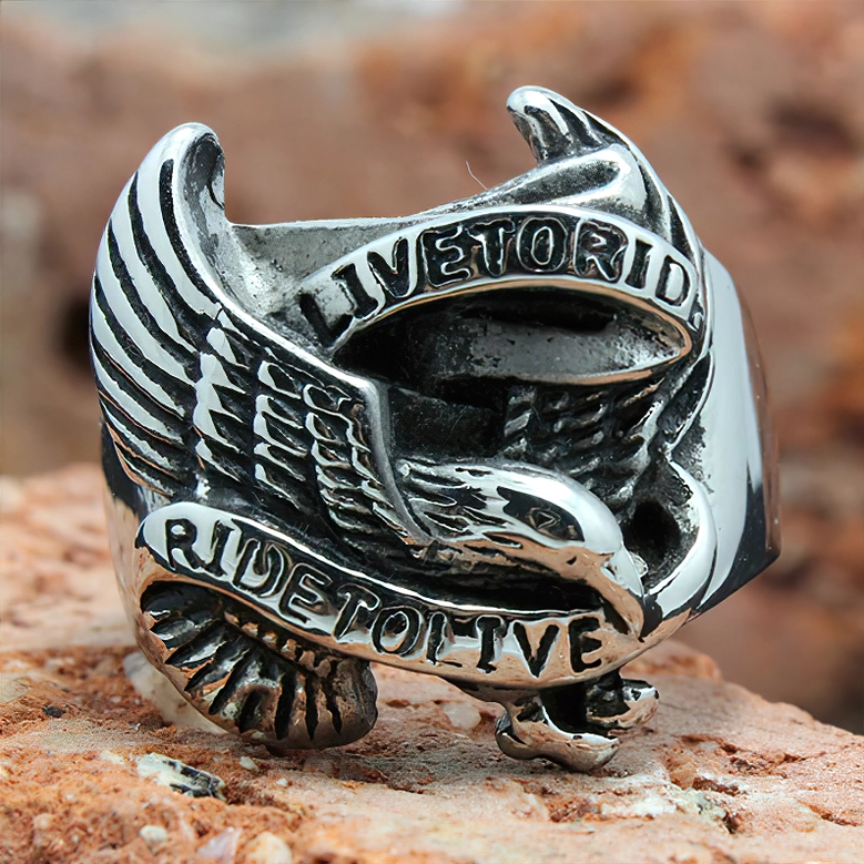 Retro gothic eagle stainless steel biker ring close-up showing detailed feather carvings and text engravings, outdoor rocky setting