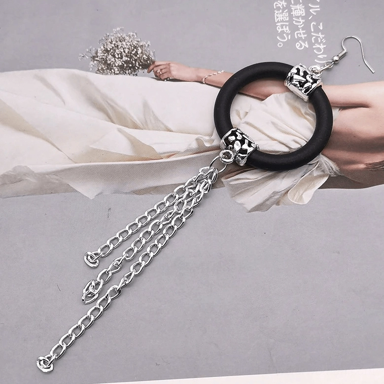 Single black rubber hoop earring with double-strand silver chain tassel on mannequin arm, flat lay with dried flowers, gothic styling