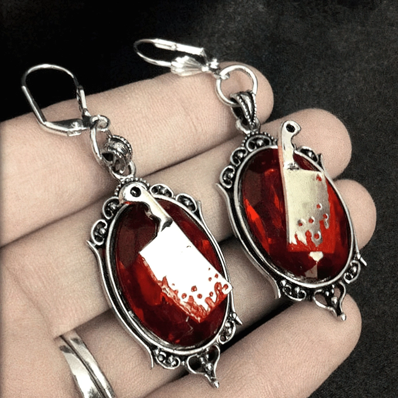 Hands-on close-up of gothic meat cleaver earrings with red gemstone and blood splatter cleaver design, hand display