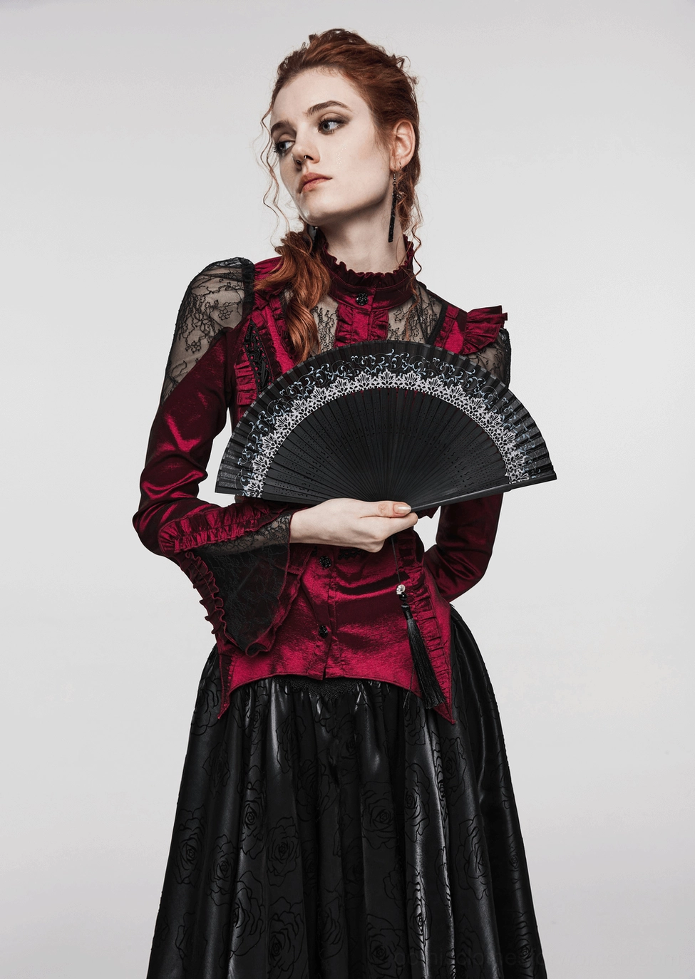 Red-haired model in red gothic lace blouse holding black hand fan, three-quarter view studio photograph