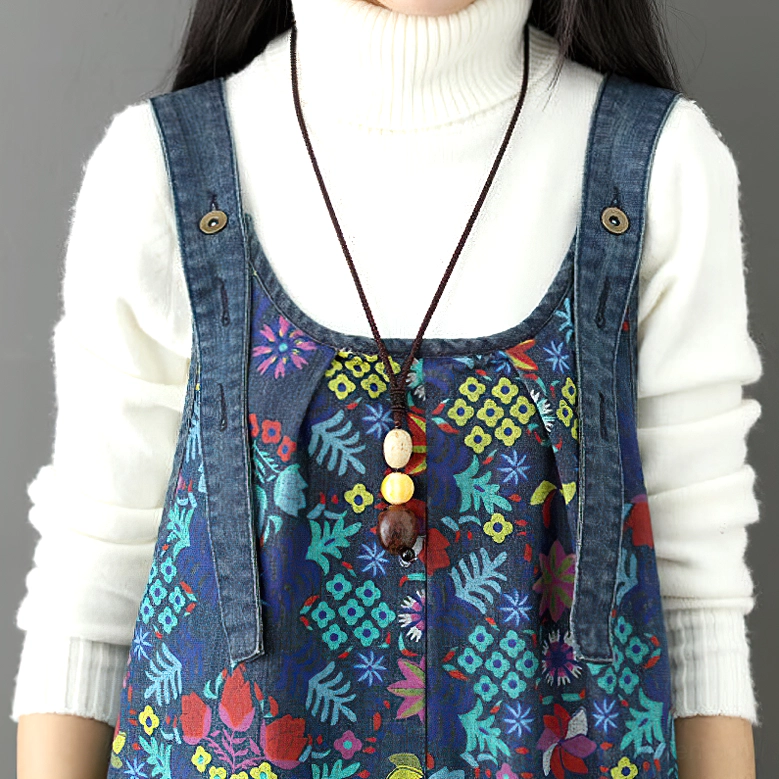 Close-up detail of floral denim overalls upper section showing buttoned straps, botanical print fabric, and beaded pendant necklace with white turtleneck