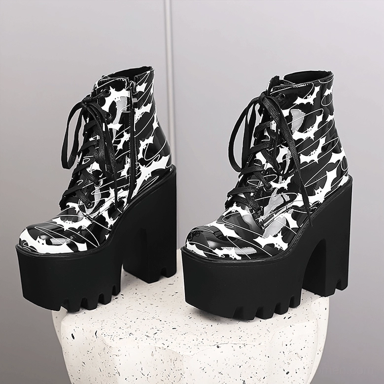 Pair of black bat print gothic platform ankle boots on white stone display stand, front three-quarter angle showing dual side zipper closures and round toe shape