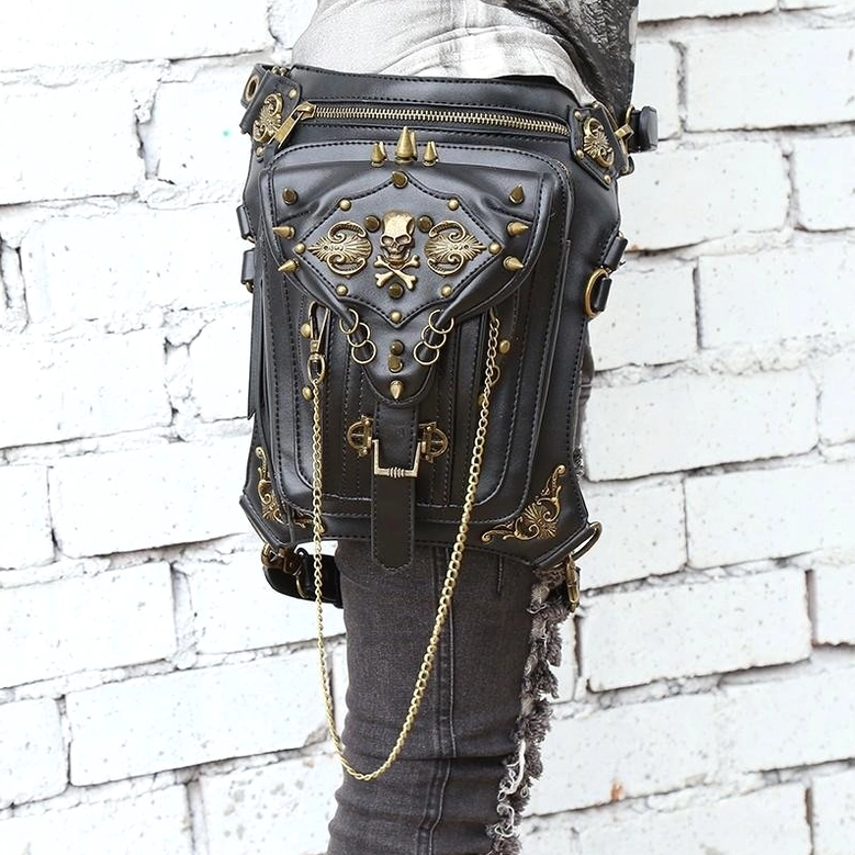 Front side view of gothic skull waist bag with bronze spike ornament and metal chains worn on dark jeans, white brick background