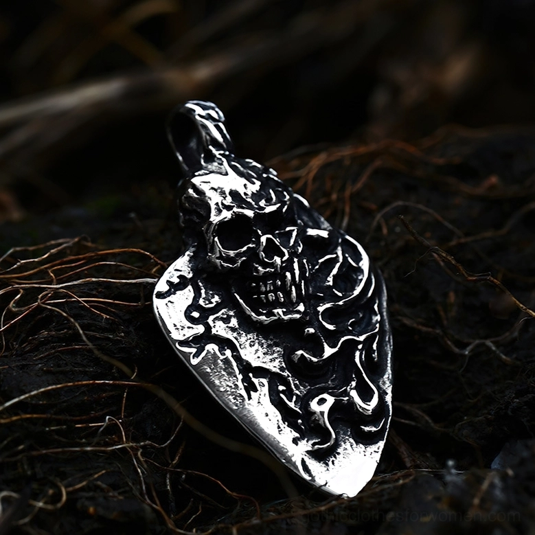 Low-angle close-up of weathered silver skull guitar pick pendant on dark twig surface background, highlighting aged metal finish and rough carved skull carving details