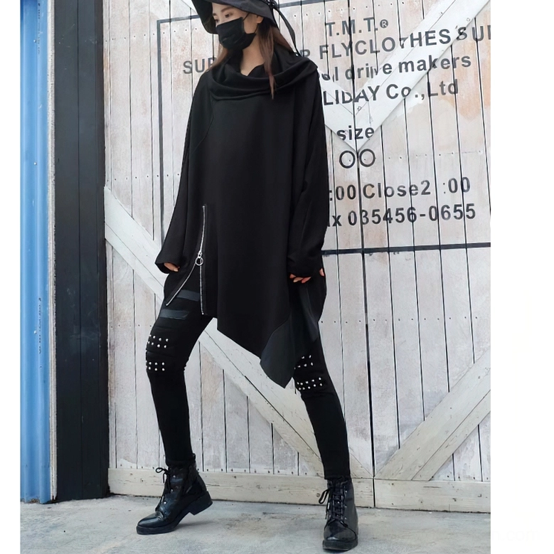 Front-side full-body shot of model in black asymmetrical gothic sweatshirt with batwing sleeves and studded pants, outdoor industrial setting