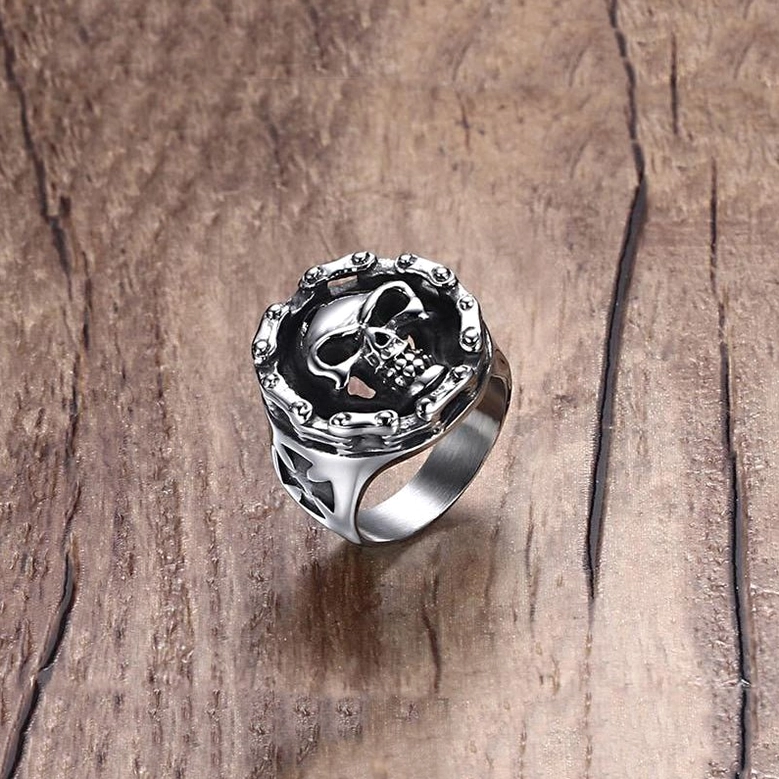 Front three-quarter perspective of alternative fashion skull ring with riveted circular frame around skull on natural wooden surface