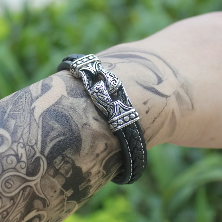 Tattooed male wrist wearing black leather gothic bracelet outdoors against green background, side view
