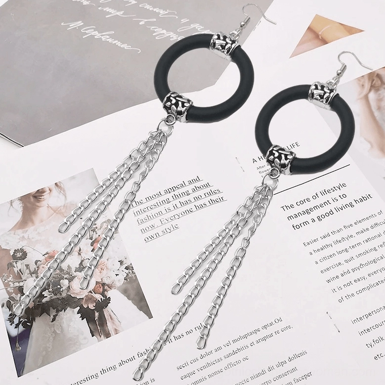 Pair of black rubber hoop drop earrings with silver chain tassels on magazine pages, overhead lifestyle display, gothic statement jewelry
