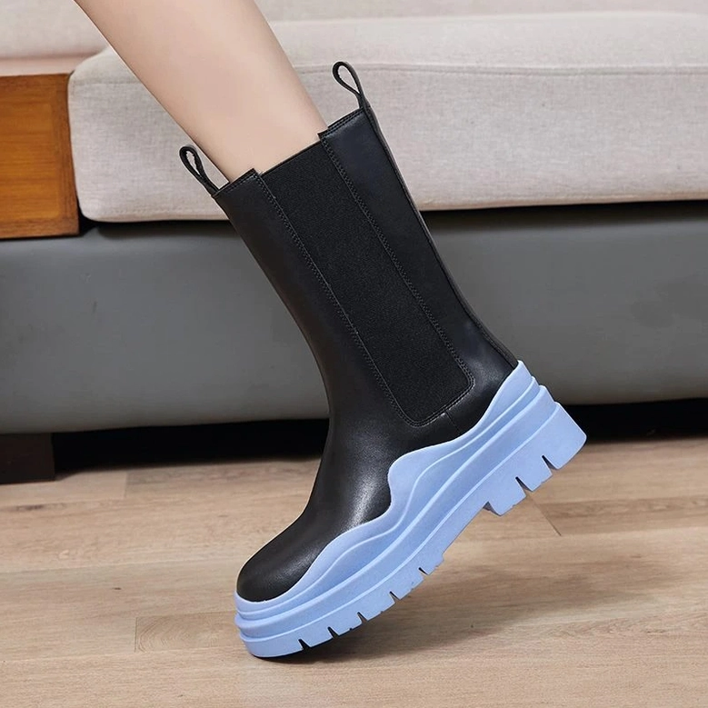 Woman model wearing black leather platform boots with blue wavy sole, front low angle on sofa background, detailed sole view