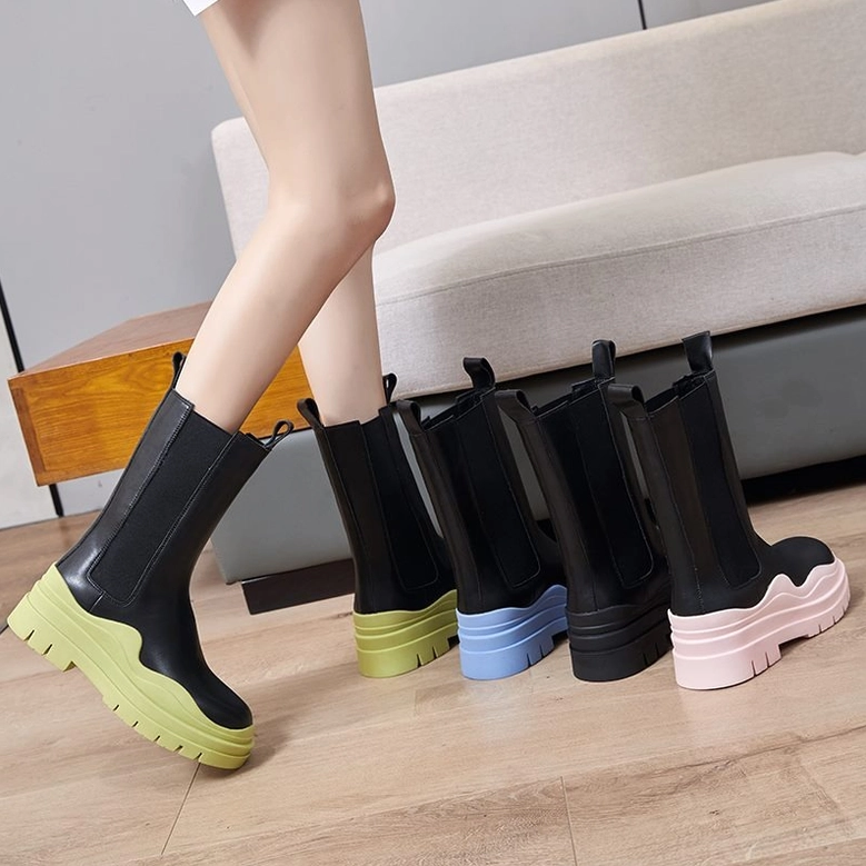 Female model wearing black leather platform boots with varied colored chunky soles, low side angle view on indoor wooden setting