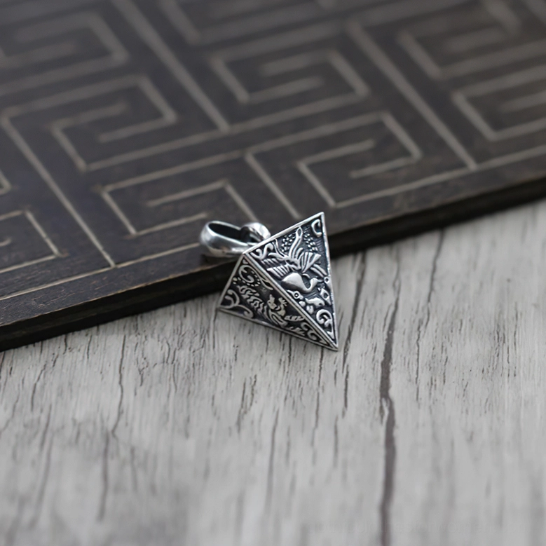Aged sterling silver pyramid pendant featuring embossed decorative motifs, side view on light wood table with dark geometric panel backdrop