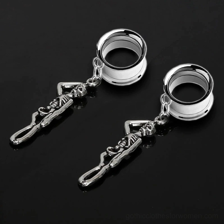 Skull ear tunnel charms lying flat on reflective black surface – stainless steel skeleton dangle earrings oblique view