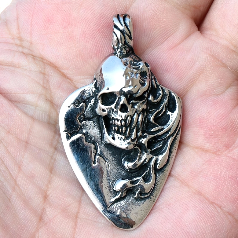 Close-up detail shot of gothic skull guitar pick pendant held in human palm, showing weathered silver finish and embossed flame textures surrounding carved skull